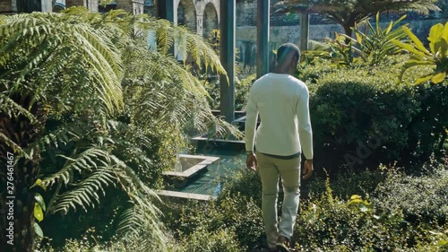 Young Black Man Model in White Wardrobe Walks Into Field With Plants in Inner City Historic Park With Pool photo