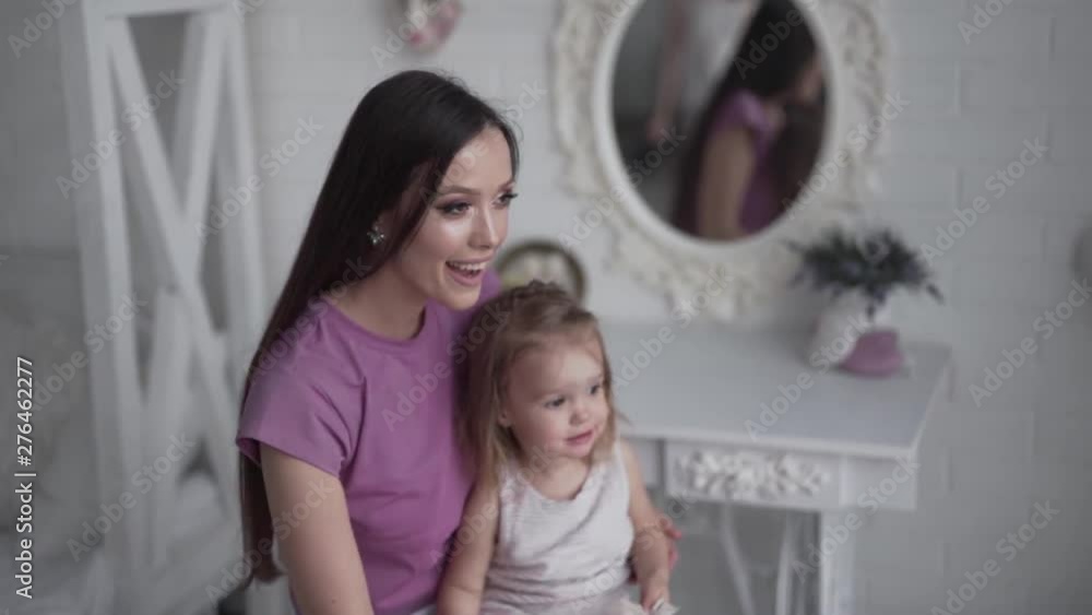 Young beautiful mother is sitting in a white-coloured room and playing with her little cute daughter. They both are wearing a rose pajamas. Young beautiful mother is sitting in a white-coloured room and playing with her little cute daughter. They both are wearing a rose pajamas.