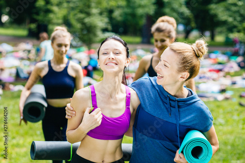 Female friends meeting for morning workout yoga or pilates exercise in summer park outdoor. Two excited women smiling to each other whilehugging and going with group people on background.