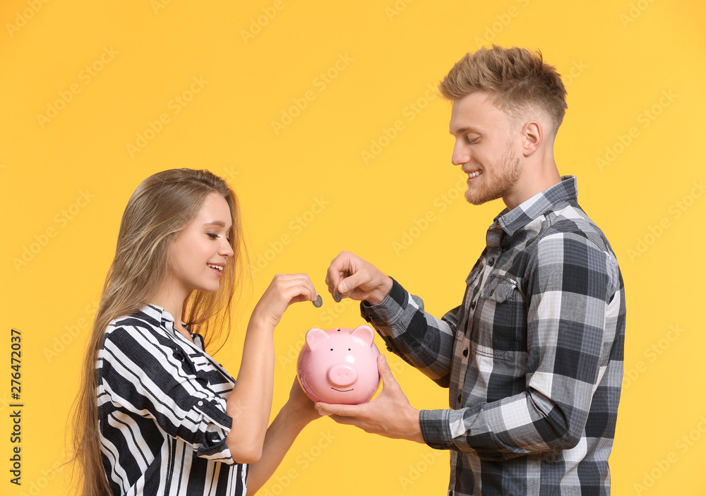 Happy couple putting coins into piggy bank on color background
