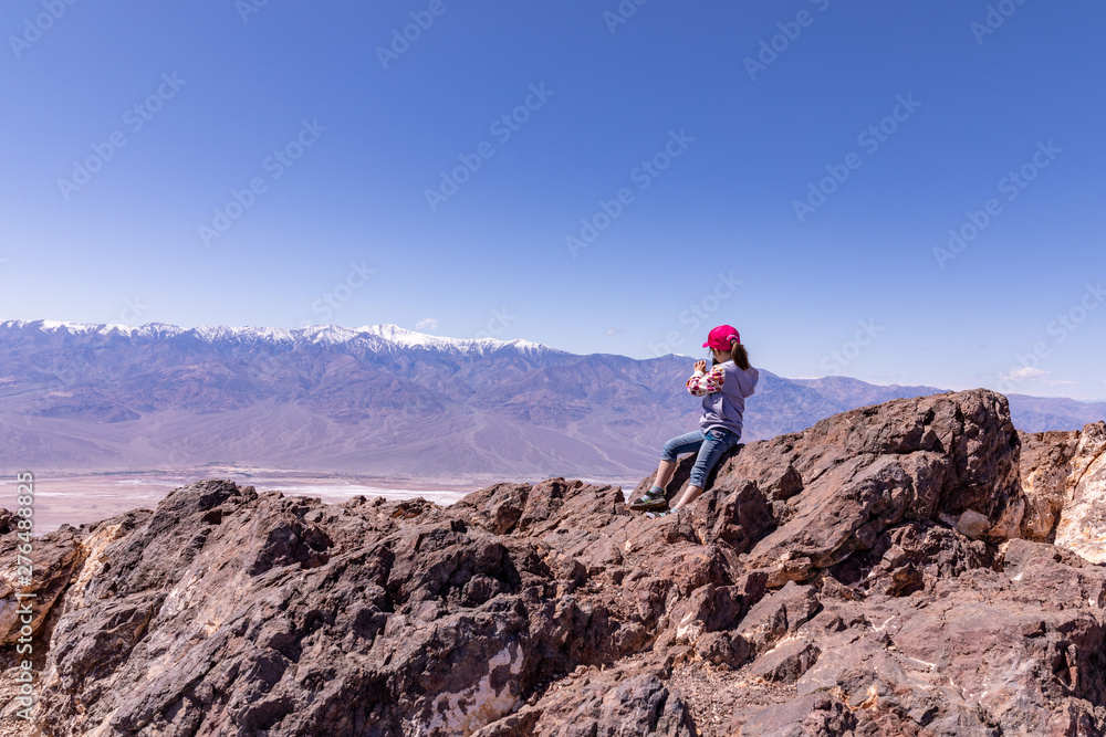 Death Valley, California \ USA - March 26 2019: Little girl in a red baseball hat, grey sweater and capri jeans taking photos of Badwater basin from rocks of Dante's viewpoint