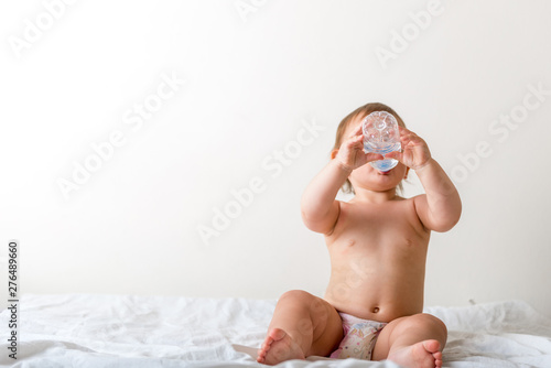 Baby toddler sits on the white bed, smiles and drinks water from plastic bottle. Copy space © troyanphoto