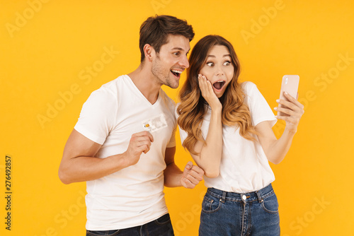 Portrait of surprised couple in basic t-shirts rejoicing while holding credit card and smartphone
