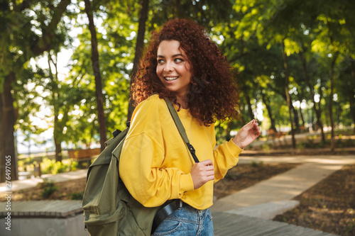 Smiling young beautiful curly student girl outdoors in nature park walking with backpack.