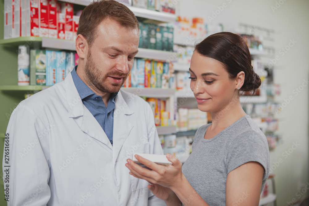 Attractive young woman talking to the chemist, shopping for medicament at the drugstore. Mature male pharmacist helping his female client