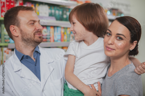 Charming young woman smiling to the camera, carrying her little son while shopping at the pharmacy. mature male chemist helping his female customer shopping for medicament with her child