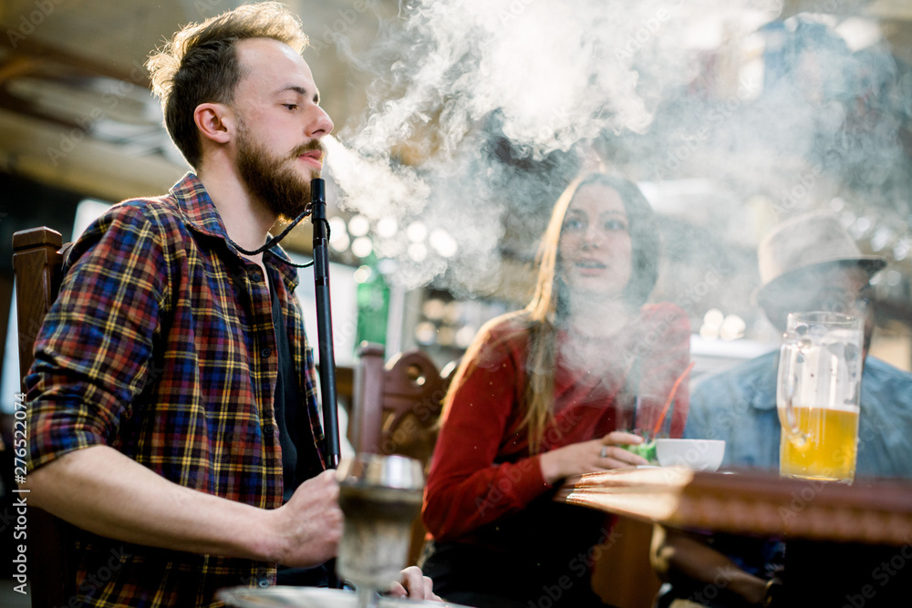 Caucasian young man in casual wear smoking a hookah while sitting at the table in cafe indoors together with friends