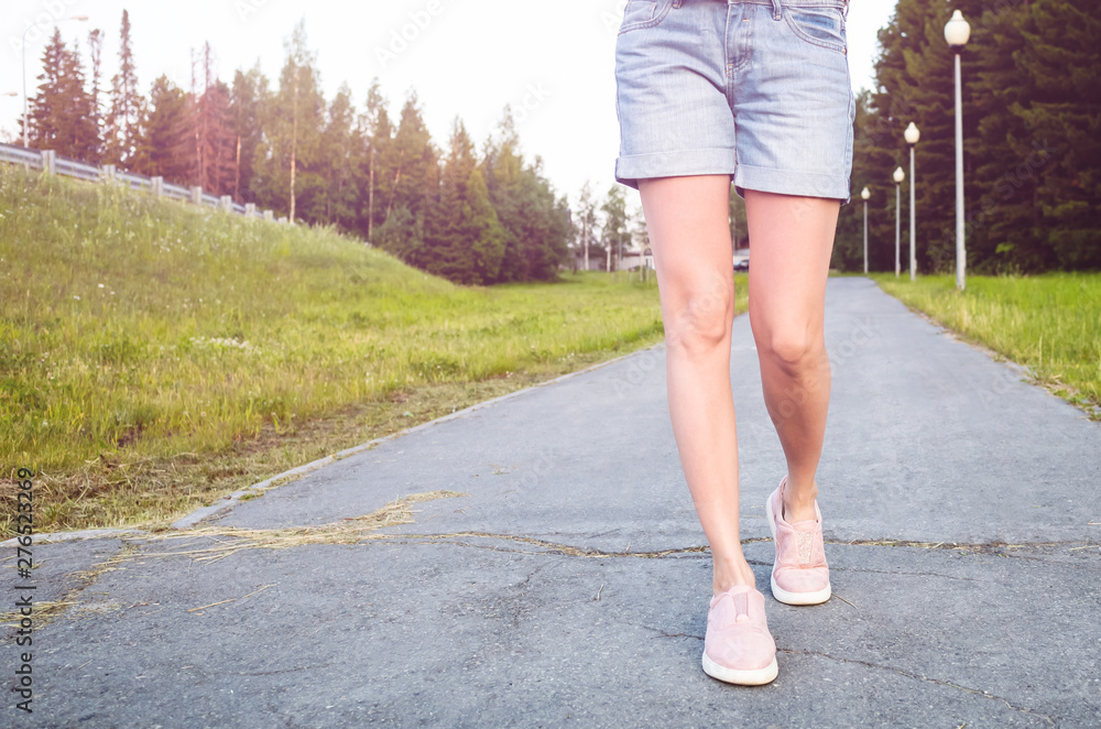 Women's slim legs in blue denim shorts and pink sneakers on the sidewalk. Behind - lights, trees, lawn. The concept of walking, a healthy lifestyle, the minimum number of steps per day. Place for text