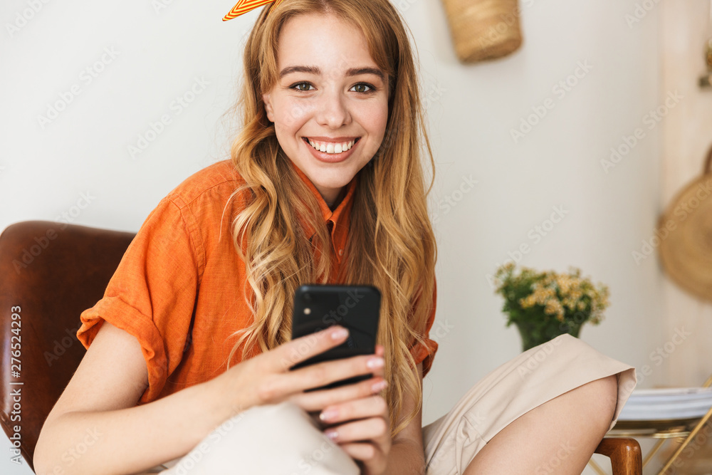 Smiling young blonde girl at home indoors using mobile phone sitting on chair. Smiling young blonde girl at home indoors using mobile phone sitting on chair.