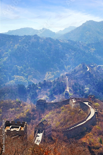 Vertical panoramic view of the Mutianyu section of the Great Wall of China, surrounded by green and yellow vegetation under a cold blue morning light.