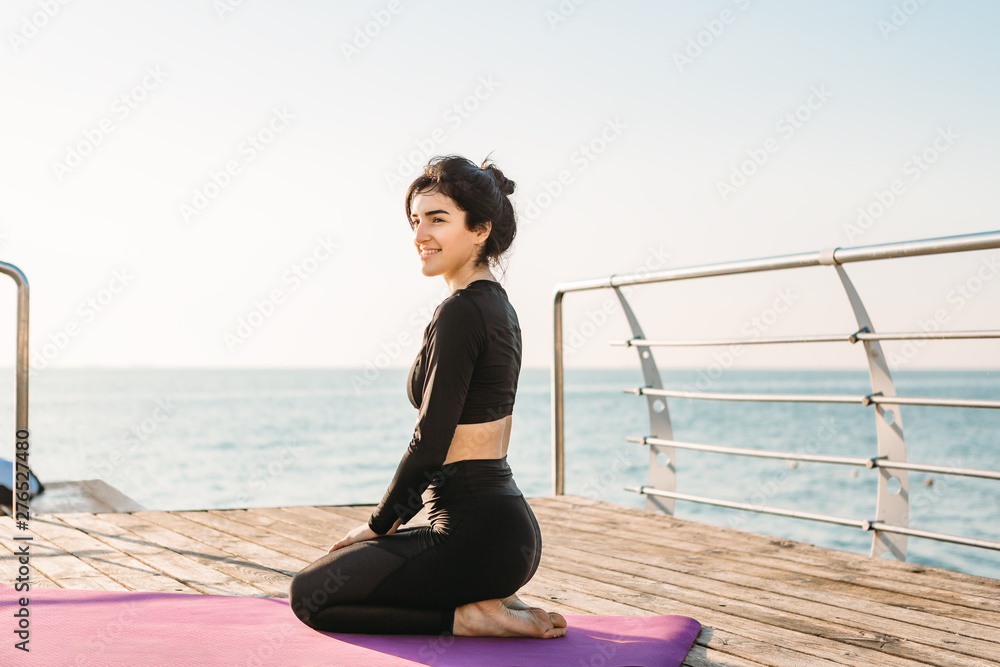 Fototapeta premium Smiling brunette young woman in a black jumpsuit rests after plank workout on a mat on pier at sunrise.