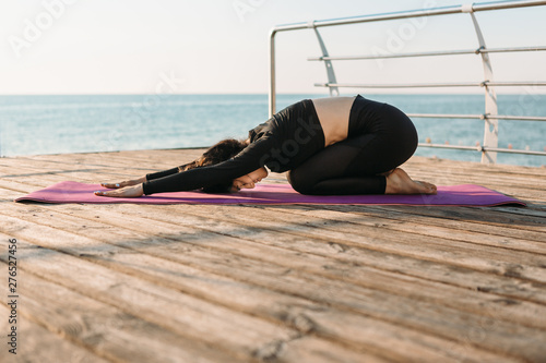 Young brunette woman in a black jumpsuit practicing yoga stretching exercises on the beach at sunrise. Concept of wellness and healthy lifestyle.