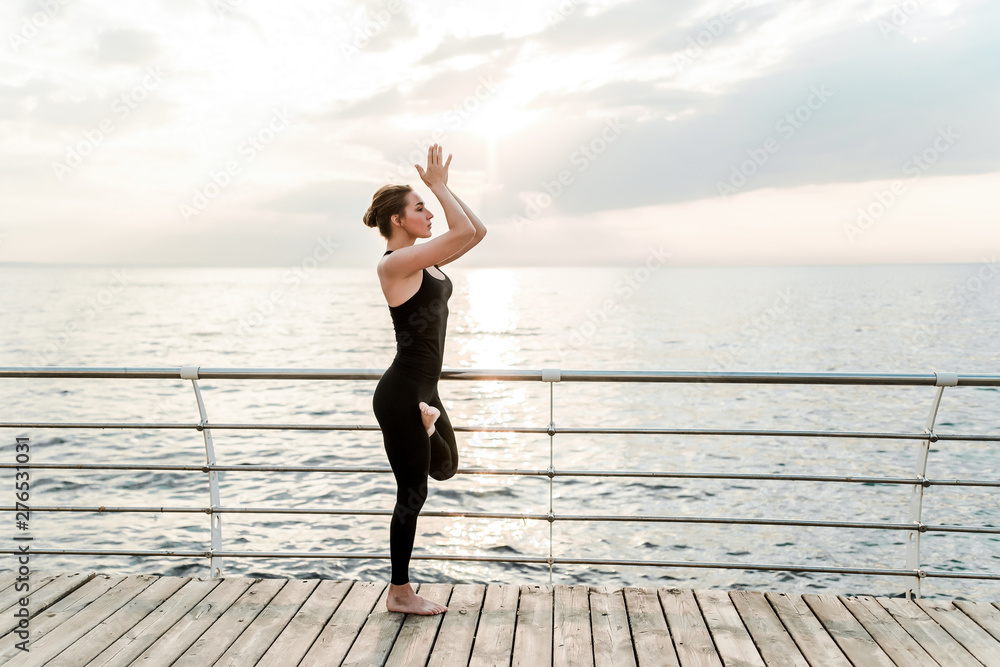 Obraz premium Flexible woman doing yoga asana near the sea on sunrise in the morning, practicing sport and fitness exercises
