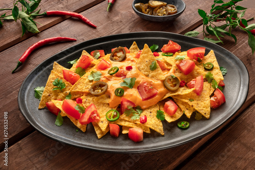 Nachos, Mexican tortilla chips, with a cheese sauce, chili and jalapeno peppers, tomatoes. and cilantro, on a dark rustic wooden background