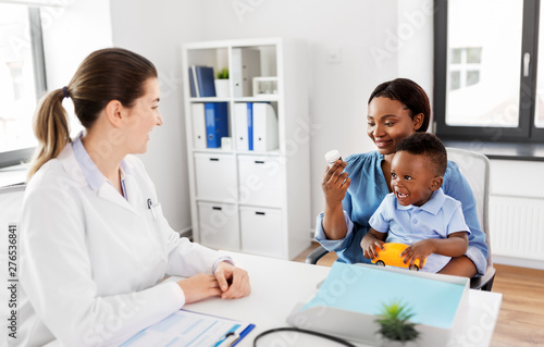 medicine, healthcare and pediatry concept - caucasian doctor and african american mother with baby son holding medication at clinic
