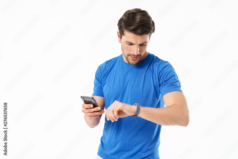 Photo of serious man in casual t-shirt looking at wristwatch and holding cellphone - obrazy, fototapety, plakaty 