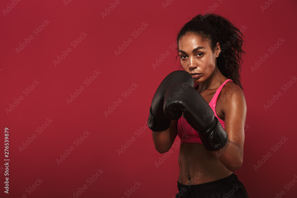 Serious strong young african sports woman boxer posing isolated over red wall background make boxing exercises.