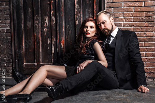 Fashion studio photo of sexy couple of sensual woman and blonde man dressed wearing classic style, sitting on the floor. Isolated on brick wall, studio shot