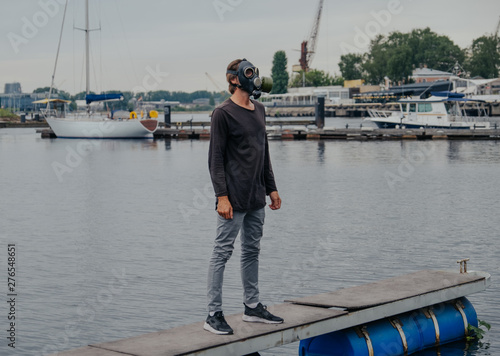 A man in a gas mask sits on a pier near the water in the city