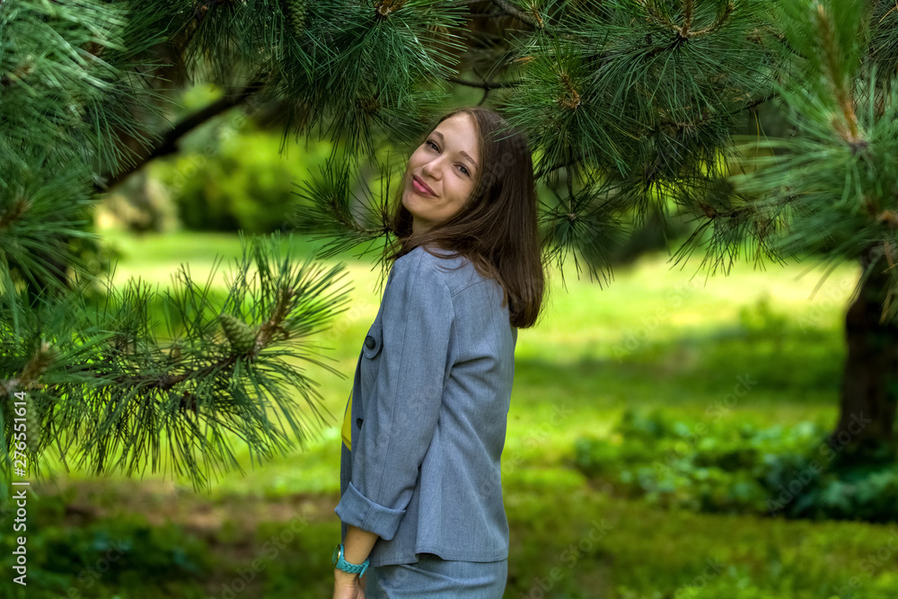 The young woman in gray business suit, in trousers among big green pines in the park. Fashion and style. Portrait. The young woman in gray business suit, in trousers among big green pines in the park. Fashion and style. Portrait.