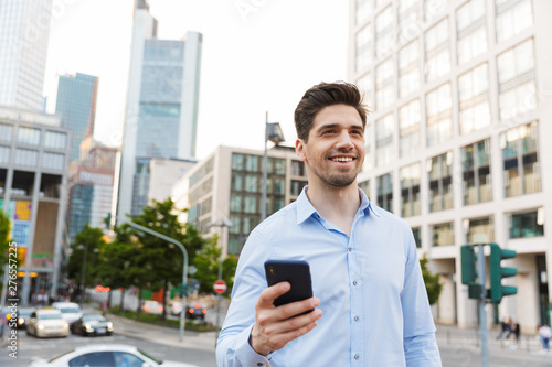 Young cheery pleased unshaved businessman walking outdoors on the street using mobile phone.