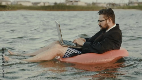 A business person is enthusiastically working behind a laptop swaying on the waves of a reservoir while sitting on a pink inflatable circle.