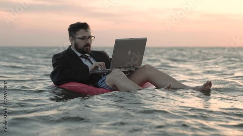 Businessman enthusiastically working behind a laptop swaying on the waves of a reservoir and sitting on a pink inflatable circle. photo
