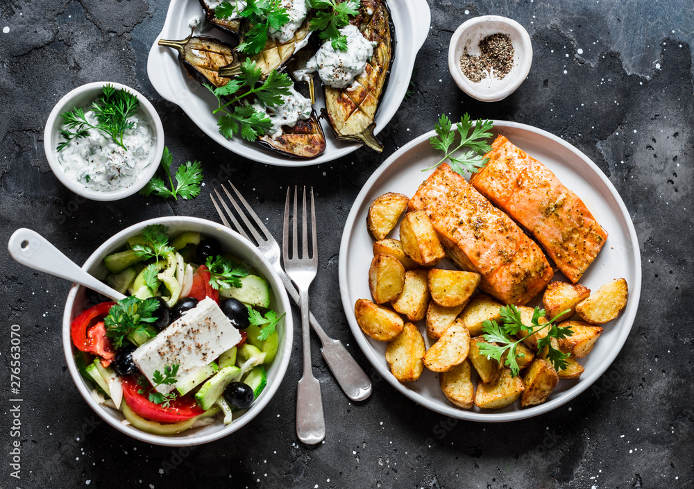 Greek style lunch table - baked lemon salmon with potatoes, greek salad, grilled eggplant with tzadziki sauce on dark background, top view. Flat lay Greek style lunch table - baked lemon salmon with potatoes, greek salad, grilled eggplant with tzadziki sauce on dark background, top view. Flat lay