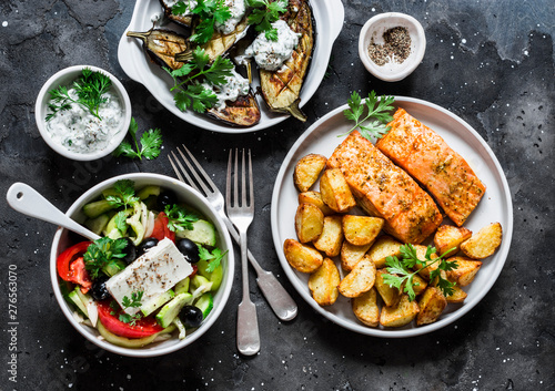 Greek style lunch table - baked lemon salmon with potatoes  greek salad  grilled eggplant with tzadziki sauce on dark background  top view. Flat lay