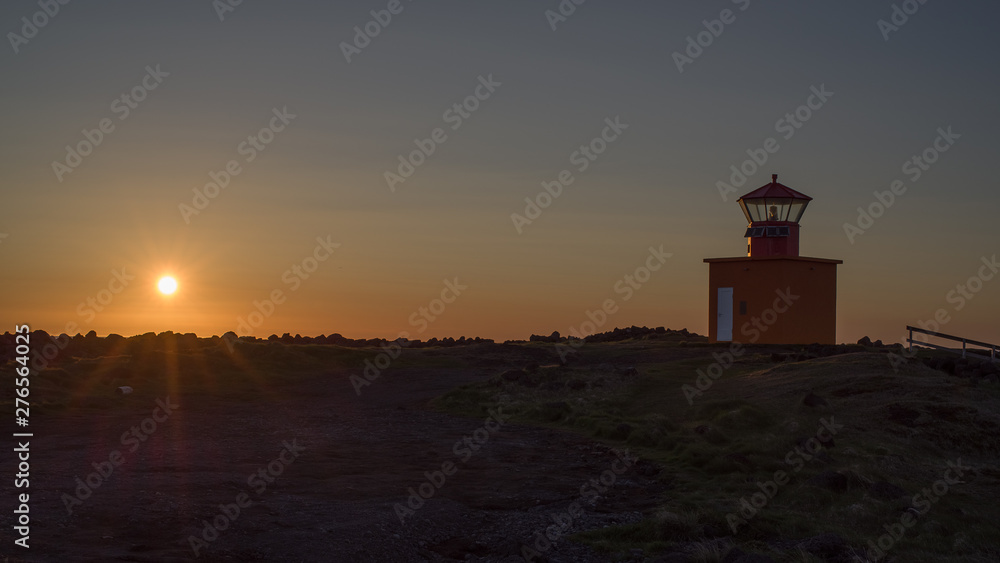 Icelandic  sunset - view of low sun above Atlantic with a lighthouse in foreground. Back light (close to midnight) throws beams on a rocky coastline dominated by a red land orange lighthouse.