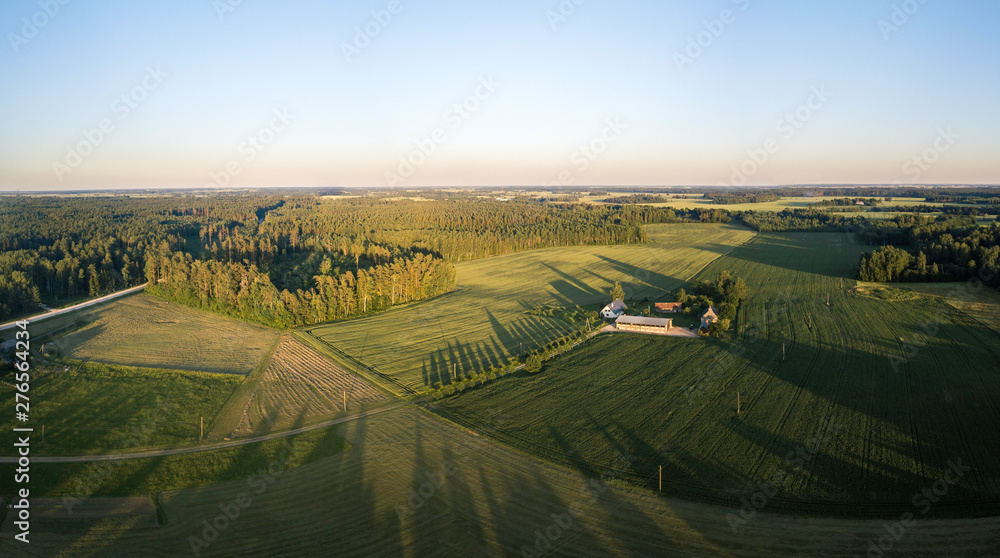 Fototapeta premium Aerial view over rural landscape in a warm summer sunset tones. Agriculture land mixing with forest and meadows. Green crop fields along the curved river. Trees creating long shadows. 