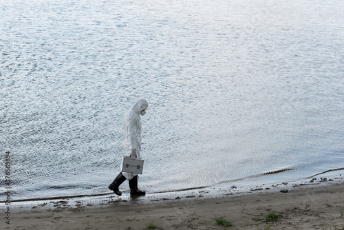 full length view of water inspector in protective costume and respirator holding inspection kit while walking on river coast