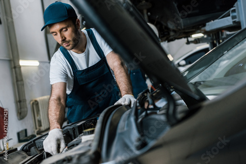 selective focus of handsome car mechanic in gloves looking at car engine