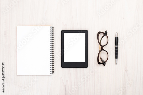 Flat lay E-reader, paper notepad, glasses and pen on wooden surface. Top view woman accessories, feminine desk with e-book and stationary stuff. Female office desktop concept. © photoguns