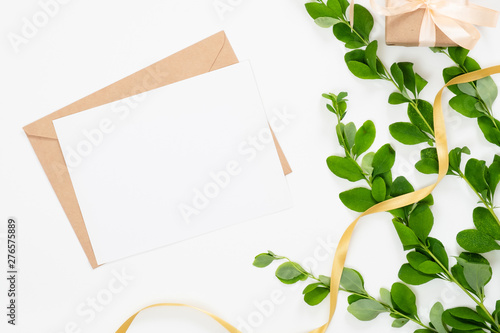 Wedding invitation card, craft paper envelope, ribbon and branch with green leaves on white background. Minimal flat lay style composition, top view, overhead.