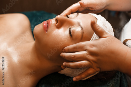 Side view portrait of a beautiful young caucasian woman leaning on a spa bed with closed eyes having facial massage in a wellness spa center.