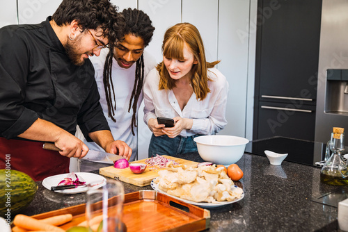 Obraz na płótnie Professional cook teaching a black guy and a red Caucasian girl how to cook. She carries her mobile in her hand to make a video. Modern kitchen. w salonie