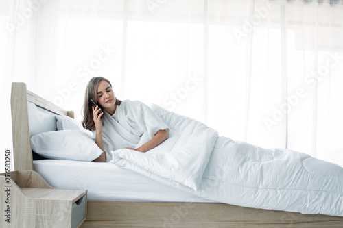 Woman talking smartphone on bed with a smiling face after waking up.