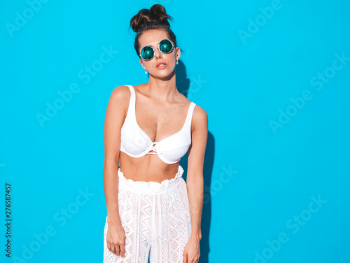 Young beautiful sexy woman with ghoul hairstyle. Trendy girl in casual summer white pants and bra in sunglasses. Hot model isolated on blue