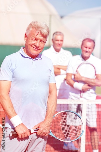 Portrait of confident mature man holding tennis racket while standing against friends on court during sunny day