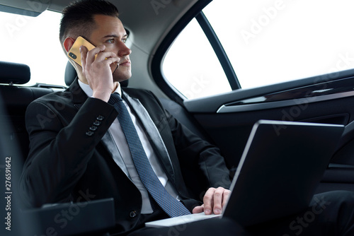 Businessman sitting in car, dressed in suit and tie. Working on laptop and talking on mobile phone. © less.talk