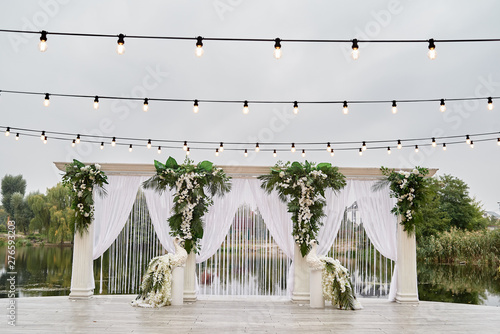 Luxury wedding arch decorated with palm leaves, orchid flowers, floral peacocks and bulbs garland outdoors, copy space. Wedding ceremony near water in park
