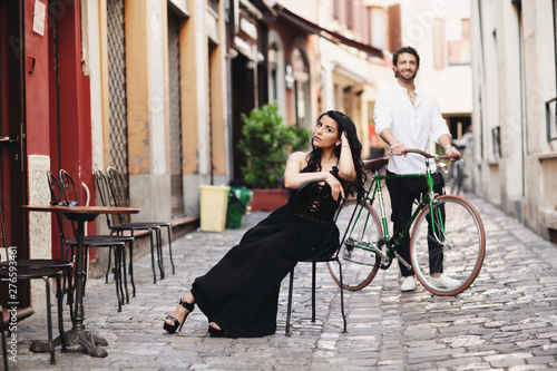 A loving couple in the old city. A woman in a black dress sits on a chair. The man behind her stands with a green bike