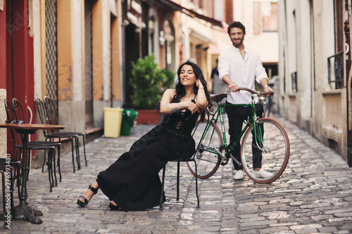 A loving couple in the old city. A woman in a black dress sits on a chair. The man behind her stands with a green bike