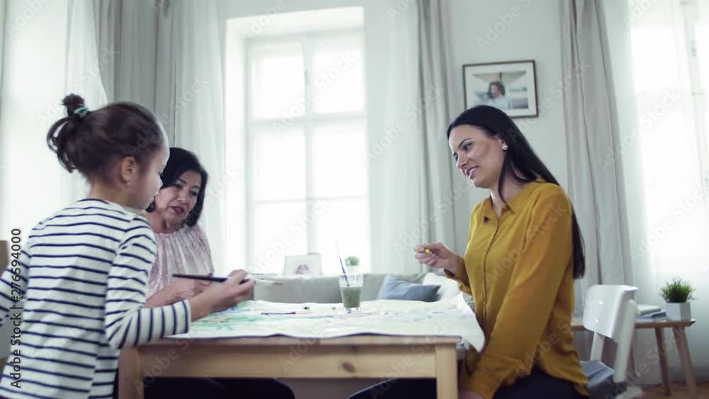 A small girl with mother and grandmother sitting at table at home, painting pictures. A small girl with mother and grandmother sitting at table at home, painting pictures.