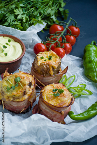 Baked new potatoes wrapped in bacon, on top cheese, microgreen, sauce and fresh vegetables. Dark background.