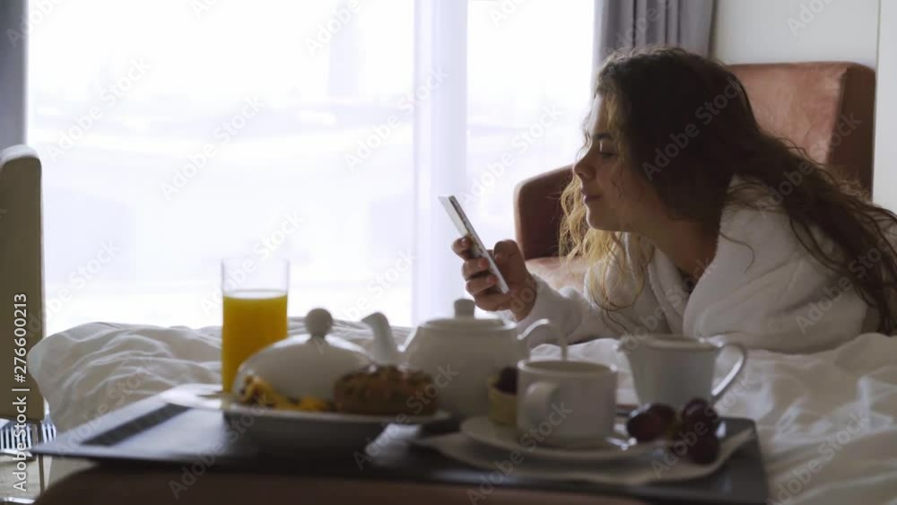 Woman have breakfast in modern hotel apartment. Close-up portrait young girl in bathrobe lying on bed using smartphone in hands. Out of focus tray with meal, juice, plate, teapot on foreground Woman have breakfast in modern hotel apartment. Close-up portrait young girl in bathrobe lying on bed using smartphone in hands. Out of focus tray with meal, juice, plate, teapot on foreground