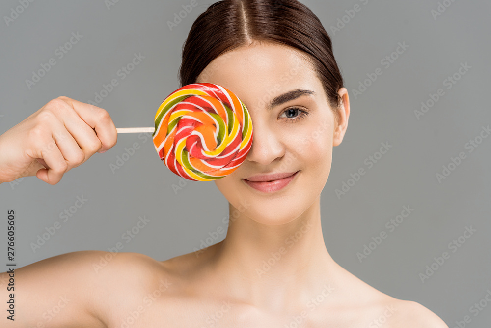 cheerful naked woman covering eye with colorful lollipop isolated on grey