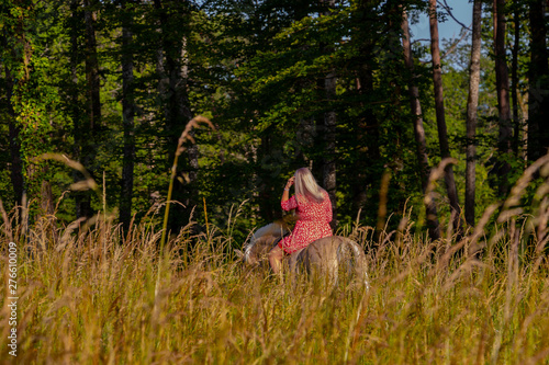 junge Frau im roten Sommerkleid reitet auf einem Pferd neben dem Wald und einem Weizenfeld bei wunderschönem Sommerwetter