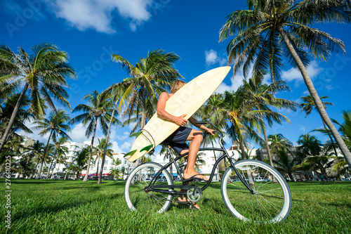 Surfer with blond hair carrying his surfboard on a beach cruiser bike through a tropical palm tree landscape in Miami, Florida, USA